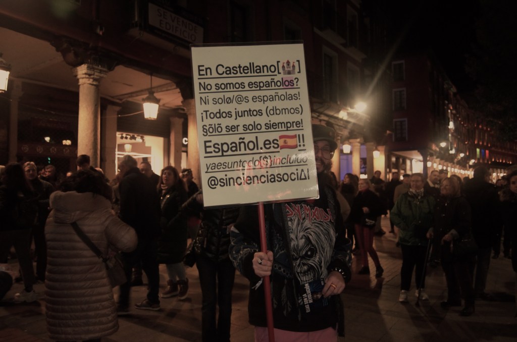 Manifestante muestra un cartel con el texto "En castellano no somos españolos? Ni solo españolas! ¡Todos juntos (dbmos) solo ser solo siempre! Español.es!! (yaesuntodoincluido) @sincienciasocial"